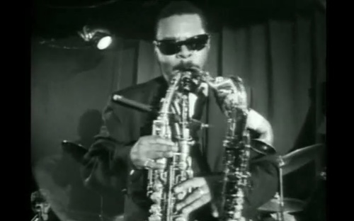 A man in sunglasses passionately plays two saxophones simultaneously on stage. The black-and-white image captures an intense, soulful jazz performance.